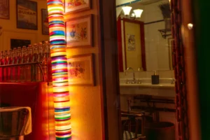 Marc · 20/12/2025 · 6045 x 4030 Cette photographie montre l'intérieur d'un bar ou d'un restaurant. Le sujet principal est une colonne décorée de bandes lumineuses colorées (orange, rouge, jaune), éclairée de façon dramatique. La composition met en valeur cette colonne centrale, encadrée par des tables et des chaises en bois.Le cadrage est serré, créant une ambiance intimiste. La lumière est tamisée et chaude, avec des jeux d'ombres qui accentuent l'atmosphère cosy. Les couleurs dominantes sont les tons chauds (orange, rouge, brun), renforçant une ambiance chaleureuse et conviviale, typique d'un lieu de détente.