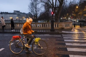 Benoit · 20/12/2025 · 6910 x 4607 Cette photographie capture une personne à vélo, sujet principal, vêtue d'une veste orange et tenant un bouquet jaune. La composition montre une rue urbaine avec des bâtiments et des arbres en arrière-plan.Le cadrage est serré sur le cycliste, mais inclut aussi des éléments architecturaux et urbains. La lumière, tamisée et douce, suggère un crépuscule ou une soirée d'hiver. Les couleurs dominantes sont les tons froids (gris, bleu nuit) adoucis par les éclairages artificiels et les touches chaudes (orange, jaune).L'ambiance est calme et mélancolique, évoquant une promenade solitaire en ville. Le style est réaliste et documentaire.