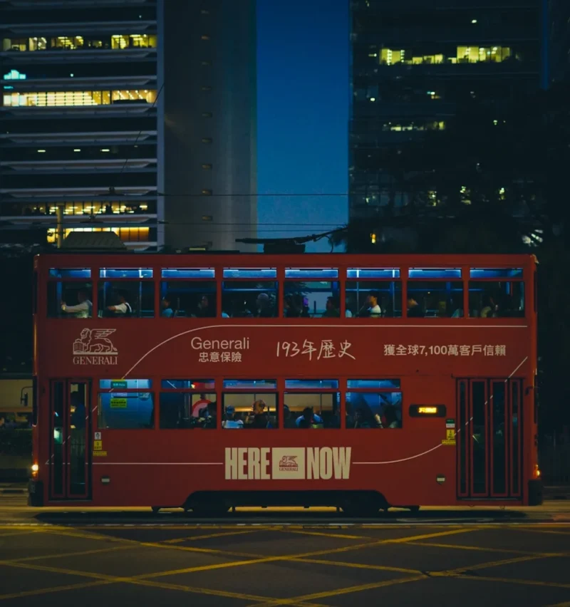 Romain · 23/01/2018 · 1016 x 1082 Cette photographie met en scène un **double-decker bus rouge** comme sujet principal, marqué d’une publicité lumineuse "HERE AND NOW". La composition est centrée sur le bus, avec un cadrage serré et légèrement bas, accentuant sa taille imposante. L’arrière-plan montre des **gratte-ciel éclairés**, créant un contraste entre l’architecture moderne et le bus vintage.Les **couleurs dominantes** sont le rouge vif du bus et les tons bleus/blancs des lumières urbaines. La lumière, nocturne et artificielle, donne une ambiance **dynamique et énergique**, typique d’une ville animée. Le style est **urbain et contemporain**, mêlant nostalgie et modernité.