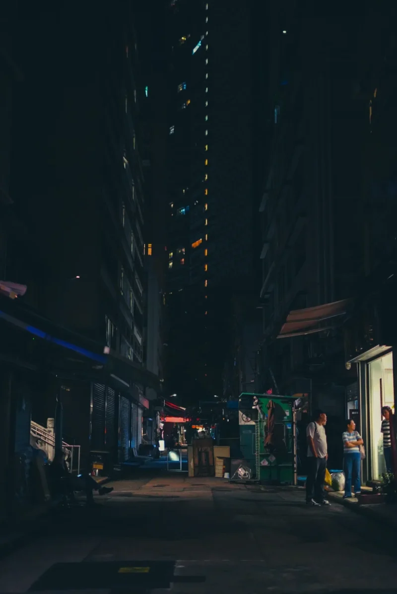 Romain · 23/01/2018 · 1080 x 1616 Cette photographie capture une rue urbaine nocturne. Le **sujet principal** semble être l’allée sombre entre des bâtiments éclairés par des néons et des enseignes.La **composition** est symétrique, avec des lignes verticales des immeubles convergeant vers le lointain. Le **cadrage** est serré, créant une perspective profonde.La **lumière** est faible et artificielle, avec des éclairages bleutés et jaunes, donnant un contraste marqué. Les **couleurs dominantes** sont des teintes sombres et froides, avec des touches de bleu et de jaune.Le **style** est réaliste et légèrement flou, évoquant une ambiance **mélancolique et mystérieuse**.