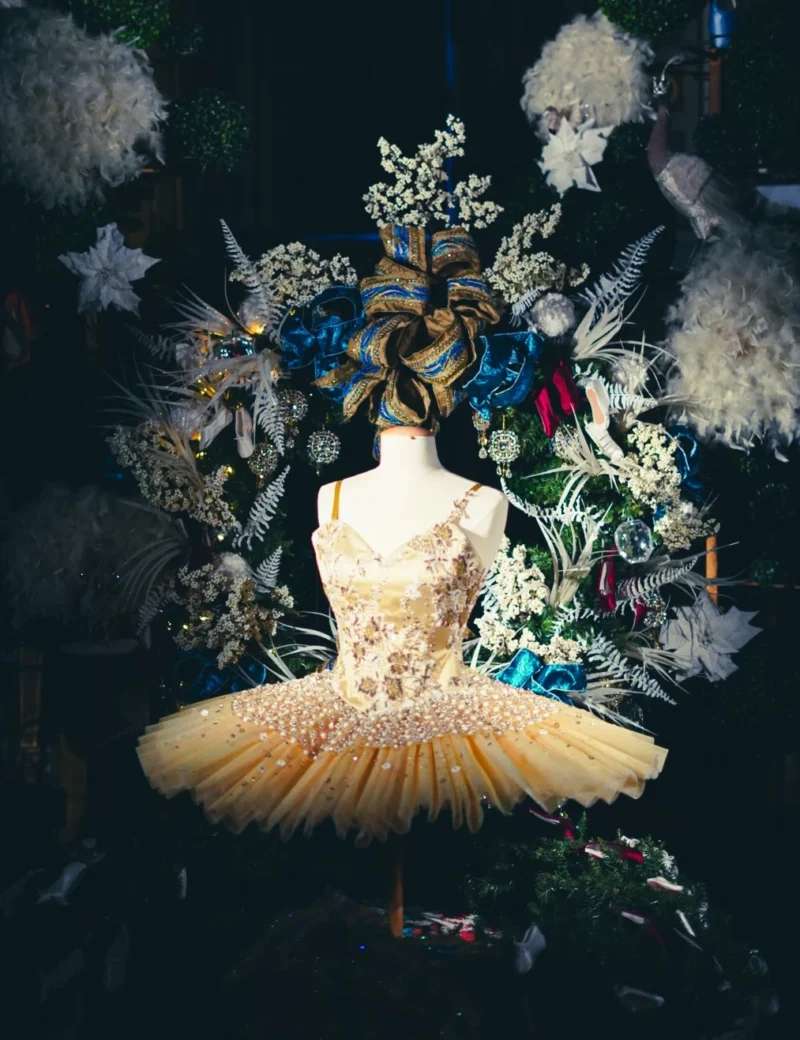 Romain · 07/02/2018 · 1013 x 1317 Cette photographie met en scène une silhouette féminine en costume de ballet élégant, aux tons clairs et à la jupe volumineuse. La composition est symétrique, avec un cadrage serré centré sur le sujet, entouré d’une profusion de fleurs et de feuillages artificiels colorés.La lumière douce et tamisée accentue les détails et crée une ambiance onirique. Les couleurs dominantes sont les blancs, jaunes pâles et touches de bleu et vert. Le style évoque un mélange de classicisme et de fantaisie baroque, renforcé par l’arrière-plan luxuriant. L’ambiance est à la fois mystérieuse et poétique, presque théâtrale.
