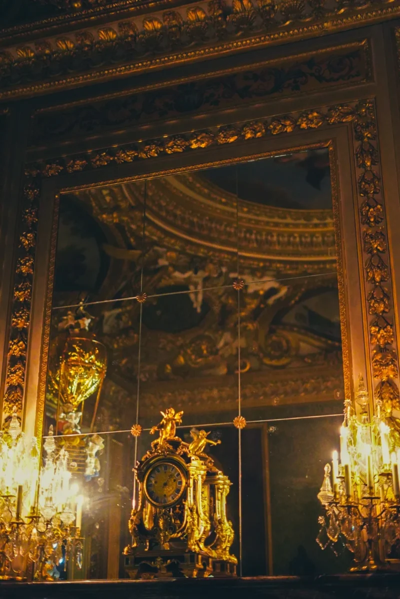 Romain · 07/02/2018 · 1080 x 1616 Cette photographie montre un miroir orné encadré de motifs dorés et sculptés, reflétant une décoration intérieure riche. Le sujet principal est le cadre luxueux et les chandeliers éclairés de chaque côté.La composition est symétrique, avec un cadrage serré mettant en valeur les détails dorés. La lumière, chaude et tamisée, provient des bougies, créant une ambiance intimiste et élégante. Les couleurs dominantes sont les tons dorés et bruns, renforçant un style baroque ou classique. L’ambiance est opulente et raffinée, évoquant le luxe et le prestige.