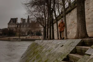 Ahmet · 20/12/2025 · 2500 x 1665 Cette photographie met en scène un homme en manteau marron comme sujet principal, debout près d’un quai en pierre. La composition joue sur la perspective avec des marches descendant vers un cours d’eau.Le cadrage est serré sur l’homme, encadré par des arbres et un bâtiment historique en arrière-plan. La lumière est douce et diffuse, typique d’un jour gris et froid. Les couleurs dominantes sont les tons neutres (gris, marron, beige) avec des touches de vert sur les pierres et les arbres.Le style est réaliste et sobre, évoquant une ambiance mélancolique et contemplative.