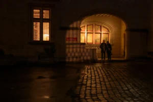 Ahmet · 20/12/2025 · 2500 x 1667 Cette photographie capture deux silhouettes floues près d'une entrée éclairée, probablement des personnes discutant. La composition repose sur un cadrage serré, mettant en valeur l’architecture et les jeux d’ombre. La lumière douce et chaude, filtrée par les fenêtres, crée une ambiance tamisée et intimiste. Les couleurs dominantes sont les tons chauds (jaunes, oranges) contrastant avec les teintes sombres de la nuit.Le style est réaliste et légèrement artistique, avec un flou artistique qui accentue l’atmosphère mystérieuse et poétique. L’ambiance évoque la sérénité et une touche de mélancolie.