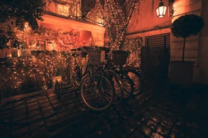 Ahmet · 20/12/2025 · 2500 x 1667 Cette photographie met en scène un vélo attaché à un support devant une façade illuminée. Le sujet principal est le vélo, avec ses accessoires (panier, lumière).La composition est simple et symétrique, avec une perspective légèrement basse. Le cadrage est serré, centré sur le vélo et son environnement immédiat.Les couleurs dominantes sont des tons chauds et orangés, créant une ambiance tamisée et chaleureuse. La lumière douce, probablement artificielle, accentue les ombres et donne un style rétro ou nostalgique.L’ambiance est paisible et intimiste, évoquant une soirée calme en ville.