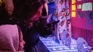 Cette photographie montre deux personnes jouant à un jeu vidéo rétro, probablement un flipper ou un jeu d'arcade. Le sujet principal est la personne en premier plan, concentrée sur le jeu.La composition est dynamique, avec un cadrage serré sur les joueurs et l’écran lumineux. Les couleurs dominantes sont vives : bleu électrique, rouge et jaune, créant une ambiance rétro et ludique. La lumière est artificielle et colorée, renforçant l’aspect festif et nostalgique. Le style évoque les salles d’arcade des années 80-90, avec une ambiance joyeuse et immersive.