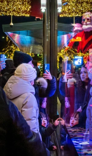 Cette photographie capture un groupe de personnes en extérieur, probablement lors d’une fête de nuit. Le sujet principal semble être une personne en train de prendre une photo avec son téléphone, entourée d’autres individus également occupés avec leurs appareils.La composition est dynamique, avec un cadrage serré sur les visages et les gestes. Les lumières colorées et les décorations festives (guirlandes lumineuses) créent une ambiance joyeuse et animée. Les couleurs dominantes sont les teintes vives des lumières (bleu, rouge, vert) et les tons neutres des vêtements. Le style est spontané et naturel, reflétant une ambiance festive et conviviale.