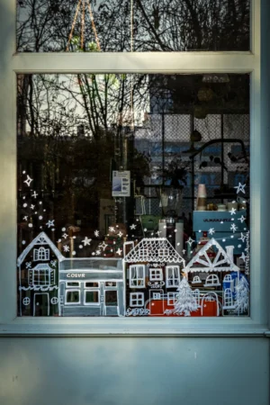 Cette photographie montre une décoration de fenêtre représentant une scène de village hivernal. Le sujet principal est une illustration de maisons enneigées avec des détails festifs, comme des guirlandes lumineuses.La composition est symétrique et bien cadrée, avec une vue à travers une vitre. La lumière, douce et naturelle, provient probablement de l'extérieur, créant des reflets et des contrastes. Les couleurs dominantes sont les tons froids (blanc, bleu) et quelques touches chaudes (rouge, orange).Le style évoque une ambiance chaleureuse et festive, typique des décorations de Noël. L'ensemble dégage une ambiance magique et réconfortante.