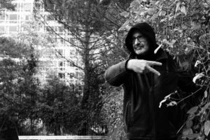 Deniz · 06/12/2025 · 3603 x 2402 Cette photographie en noir et blanc met en scène un homme comme sujet principal, vêtu d’un hoodie et tenant une cigarette. La composition est simple, avec un cadrage serré qui isole le visage et le haut du corps. L’arrière-plan, flou, révèle des arbres et un bâtiment, suggérant un cadre urbain.La lumière semble naturelle, douce mais contrastée, accentuant les ombres et les traits du visage. Le style est documentaire et brut, évoquant une ambiance intime et légèrement mélancolique. Les tons dominants, en niveaux de gris, renforcent une atmosphère sobre et contemplative.