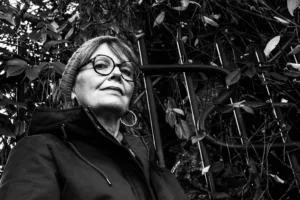 Deniz · 06/12/2025 · 3273 x 2182 Cette photographie en noir et blanc met en scène une femme comme sujet principal, vêtue d’un manteau sombre et portant des lunettes rondes. Le cadrage est serré, centré sur son visage et sa silhouette, avec un arrière-plan flou de feuilles et branches, suggérant une forêt ou un jardin.La lumière douce et naturelle crée des contrastes subtils, accentuant les traits de son visage. L’ambiance est introspective et calme, renforcée par les tons sombres et les jeux d’ombres. Le style est sobre et artistique, évoquant une atmosphère contemplative et presque mélancolique.