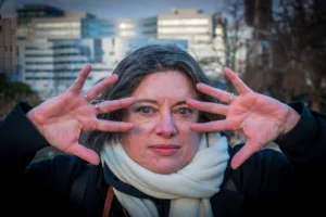 Deniz · 06/12/2025 · 6000 x 4000 Cette photographie met en scène une femme comme sujet principal, dont le visage est partiellement caché par ses mains en forme de cornes. Le cadrage est serré sur son visage, avec un arrière-plan flou de bâtiments urbains.La lumière semble naturelle, probablement en journée, avec des contrastes doux. Les couleurs dominantes sont les tons neutres (gris, beige) de ses vêtements et de son écharpe, rehaussés par le rouge de ses ongles et ses lèvres.Le style est réaliste et spontané, évoquant une ambiance à la fois humoristique et mystérieuse, renforcée par l'expression faciale et le jeu de mains.