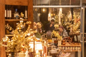 Deniz · 20/12/2025 · 2048 x 1365 Cette photographie montre un intérieur chaleureux d’une boutique artisanale ou d’un marché de Noël. Le **sujet principal** est une vendeuse derrière un comptoir décoré de **bougies, plantes séchées et décorations dorées**.La **composition** est symétrique, avec des étagères en bois remplies de produits et des suspensions lumineuses. Le **cadrage** est serré, mettant en valeur les détails et créant une ambiance intimiste. La **lumière** est douce et tamisée, renforçant l’aspect chaleureux. Les **couleurs dominantes** sont les tons dorés, verts et bois naturels.Le **style** est rustique et élégant, évoquant une **ambiance conviviale et festive**.