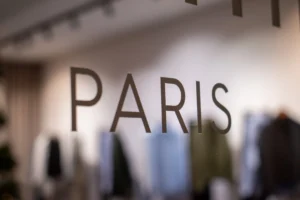 Deniz · 20/12/2025 · 2048 x 1365 Cette photographie met en avant le mot **« PARIS »** écrit en lettres stylisées sur une surface vitrée ou métallique. Le sujet principal est donc ce mot, symbolisant probablement une boutique ou un lieu lié à la mode ou au luxe parisien.La composition est épurée, avec un cadrage serré sur le texte. L’arrière-plan flou révèle des vêtements suspendus, suggérant un environnement de magasin. La lumière est douce et naturelle, créant une ambiance élégante et sophistiquée. Les couleurs dominantes sont neutres, avec des teintes grises et blanches, renforçant un style chic et intemporel. L’ambiance est raffinée et haut de gamme.