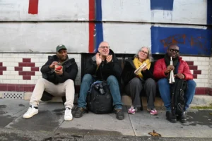 Pierre · 06/12/2025 · 6048 x 4024 Cette photographie montre quatre personnes assises sur un banc, tenant chacune un téléphone ou un objet devant leur visage. Le sujet principal est leur interaction avec leurs appareils.La composition est symétrique et serrée, avec un cadrage en plan moyen. La lumière semble naturelle, légèrement diffuse, sans ombres marquées. Les couleurs dominantes sont sobres : gris (banc, vêtements), noir et touches de rouge et bleu sur le fond mural.Le style est documentaire et spontané, évoquant une ambiance urbaine et détendue, presque intimiste malgré la présence de plusieurs personnes.