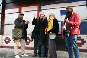 Pierre · 06/12/2025 · 5457 x 3631 Cette photographie montre quatre personnes devant un bâtiment aux couleurs vives. Le sujet principal semble être une interaction sociale, avec un homme en train de saluer ou de prendre une photo.La composition est symétrique et naturelle, avec les individus alignés verticalement. Le cadrage est serré, mettant en avant les visages et les expressions. La lumière est naturelle et semble provenir d’une source latérale, créant des ombres douces.Les couleurs dominantes sont vives : rouge, bleu, jaune et noir. Le style est documentaire et spontané, capturant un moment authentique. L’ambiance est conviviale et dynamique.