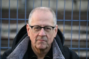 Pierre · 06/12/2025 · 6048 x 4024 Cette photographie met en scène un homme âgé comme sujet principal, au premier plan. Le cadrage est serré, centré sur son visage et ses épaules.La lumière est naturelle, légèrement contrastée, avec des ombres douces. L’arrière-plan flou, composé de barres métalliques bleutées, crée un effet de profondeur.Les couleurs dominantes sont les tons gris (vêtements, peau) et bleu froid (barres métalliques). Le style est sobre et réaliste, évoquant une ambiance sobre et introspective, presque mélancolique.