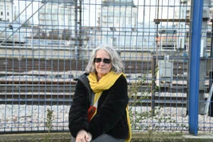 Pierre · 06/12/2025 · 6048 x 4024 Cette photographie met en scène une femme comme sujet principal, assise avec un chien. Elle porte un pull noir et un long foulard jaune, ainsi que des lunettes de soleil.La composition est simple et naturelle, avec un cadrage serré sur la femme, en avant-plan, et un arrière-plan flou de structures métalliques et de végétation. La lumière semble naturelle, probablement en journée, avec des tons doux et clairs.Les couleurs dominantes sont les tons froids (gris des structures) et chauds (jaune du foulard). L’ambiance est calme et détendue, évoquant un moment de pause dans un environnement urbain ou industriel.