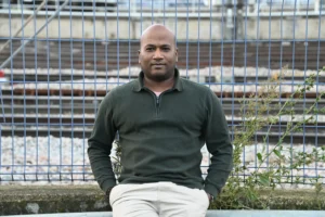 Pierre · 06/12/2025 · 6048 x 4024 Cette photographie met en scène un homme assis, sujet principal, vêtu d’un pull vert foncé et d’un pantalon clair. Le cadrage est serré, centré sur lui avec un arrière-plan flou de structure métallique et végétation.La lumière semble naturelle, douce et légèrement latérale, créant des ombres subtiles. Les couleurs dominantes sont les tons verts (pull, feuillage) et gris (structure métallique). L’ambiance est sobre et calme, avec une touche d’urbanité et de simplicité. Le style est épuré et contemporain.