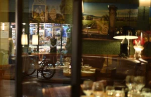 Pierre · 20/12/2025 · 4933 x 3149 Cette photographie montre un intérieur de café ou restaurant vu à travers une vitre. Le sujet principal est un vélo garé devant la vitrine, reflété partiellement.La composition est symétrique avec des éléments intérieurs (tables, chaises, luminaires) et extérieurs (bicyclette, affiches). Le cadrage est serré, centré sur la vitrine.La lumière est tamisée, créant une ambiance chaleureuse et intimiste. Les couleurs dominantes sont les tons chauds (bois, brun) et des éclairages dorés. Le style est réaliste et légèrement flou, évoquant une ambiance nocturne et cosy.