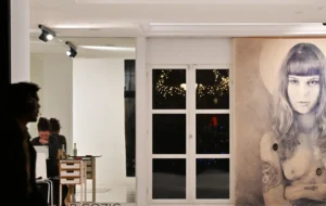 Pierre · 20/12/2025 · 3426 x 2165 Cette photographie montre une pièce moderne avec une ambiance artistique. Le sujet principal est une grande illustration murale représentant une femme stylisée aux traits marqués. La composition inclut deux personnes en discussion près d’une table basse, avec une grande fenêtre éclairée la nuit en arrière-plan.Le cadrage est large, mettant en valeur l’espace et les éléments décoratifs. La lumière est douce et tamisée, créant une ambiance chaleureuse et intimiste. Les couleurs dominantes sont neutres (blanc, gris) avec des touches sombres et des éclats de lumière naturelle. Le style est contemporain et épuré, évoquant un lieu artistique et élégant.