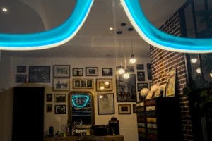 Frédéric · 20/12/2025 · 2518 x 1680 Cette photographie montre un intérieur cosy, probablement un bar ou un café. Le **sujet principal** semble être l’ambiance chaleureuse et moderne, accentuée par des **lumières LED bleutées** en forme d’arcs au plafond. La **composition** met en valeur des éléments décoratifs comme des cadres, des étagères remplies d’objets et des bouteilles.Le **cadrage** est serré, centré sur le comptoir et les détails décoratifs. Les **couleurs dominantes** sont des tons chauds (bois, brun) contrastant avec le bleu électrique des néons. Le **style** est contemporain et industriel, créant une **ambiance** à la fois intimiste et dynamique, entre convivialité et modernité.