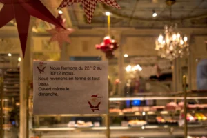 Cette photographie montre un panneau annonçant les horaires d'ouverture d'un commerce : fermé du 22/12 au 30/12 inclus, ouvert même le dimanche.Le sujet principal est ce panneau blanc avec texte noir. La composition inclut des éléments festifs en arrière-plan (décorations lumineuses, chandeliers, guirlandes). Le cadrage est serré sur le panneau, avec une lumière chaude et tamisée. Les couleurs dominantes sont les tons chauds (or, rouge, blanc), créant une ambiance chaleureuse et festive, typique des décorations de Noël.