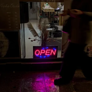 Frédéric · 20/12/2025 · 1680 x 1680 Cette photographie montre une personne devant un bar dont l’enseigne lumineuse "OPEN" est allumée sur un écran de télévision.Le cadrage est serré sur le bas du corps et l’écran. La lumière est tamisée, avec des teintes chaudes et des éclairages néon roses et bleus. Les couleurs dominantes sont des tons sombres (noir, brun) et des touches de rouge et de bleu lumineux.L’ambiance est intimiste et nocturne, évoquant un lieu convivial et moderne, peut-être un bar ou un restaurant. Le style est réaliste et spontané.