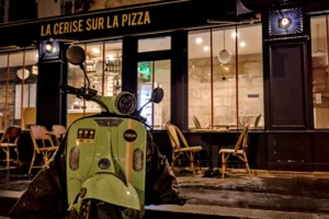 Frédéric · 20/12/2025 · 2517 x 1680 Cette photographie met en scène un scooter électrique vert fluo garé devant un restaurant nommé *« La Cerise sur la Pizza »*. La composition joue sur le contraste entre le sujet vibrant et l’arrière-plan sombre. Le cadrage serré isole le scooter, avec une légère inclinaison dynamique.Les couleurs dominantes sont le vert néon du scooter et les tons chauds des lumières intérieures du restaurant. La lumière est tamisée, créant une ambiance nocturne et mystérieuse. Le style est urbain et contemporain, évoquant une ville animée en soirée. L’atmosphère est à la fois moderne et intimiste.