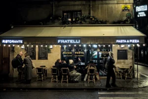 Frédéric · 20/12/2025 · 2517 x 1680 Cette photographie capture un restaurant italien nommé "Fratelli" en soirée. Le sujet principal est un groupe de personnes dînant en terrasse, éclairées par des lumières chaudes et tamisées.La composition est symétrique, avec des tables disposées en rangées. Le cadrage est serré, mettant en valeur les convives et l’ambiance conviviale. La lumière douce et artificielle crée une ambiance chaleureuse et intimiste. Les couleurs dominantes sont les tons chauds (jaunes, oranges) contrastant avec les teintes sombres de la nuit.Le style est réaliste et photographique, évoquant une ambiance nocturne romantique et détendue.