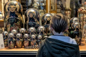 Cette photographie montre une personne observant une vitrine remplie de **poupées russes (matriochkas)**. Le **sujet principal** est le dos de cette personne, tandis que les poupées, alignées et colorées, forment un arrière-plan captivant.La **composition** est symétrique, avec un cadrage serré sur la vitrine. La **lumière** semble naturelle et douce, mettant en valeur les détails des poupées. Les **couleurs dominantes** sont vives et contrastées, typiques des motifs traditionnels russes.L’**ambiance** est à la fois intimiste et culturelle, évoquant curiosité et fascination pour l’artisanat. Le **style** est documentaire, réaliste et dépourvu d’effets stylisés.