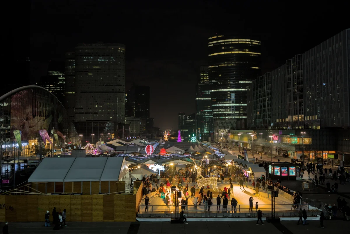 Frédéric · 14/12/2024 · 2517 x 1680 Cette photographie capture une scène nocturne animée en ville, probablement un marché de Noël. Le sujet principal est l’espace festif avec ses stands illuminés et ses visiteurs.La composition montre une perspective large, avec des bâtiments modernes en arrière-plan. Le cadrage est dynamique, incluant des éléments architecturaux et humains. Les lumières chaudes et colorées dominent, créant une ambiance conviviale et festive. Les couleurs principales sont les tons chauds (jaunes, oranges) et les éclairages bleus et violets.Le style est urbain et contemporain, évoquant une ambiance joyeuse et festive.
