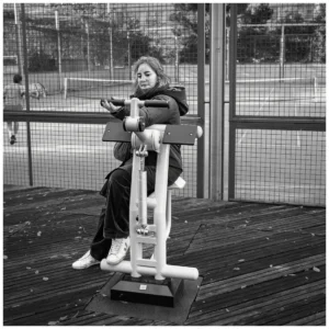 Marc Lavorata · 06/12/2025 · 1100 x 1100 Cette photographie montre une femme assise sur un banc de parc, utilisant un appareil photo ancien. Le sujet principal est centré, avec une composition simple et équilibrée.Le cadrage est serré, mettant l’accent sur elle et son activité. La lumière est naturelle, probablement en journée, avec des ombres douces. Les couleurs dominantes sont les tons gris et noirs, typiques d’une photo en noir et blanc, accentuant les contrastes.Le style est documentaire et intime, évoquant une ambiance calme et nostalgique.