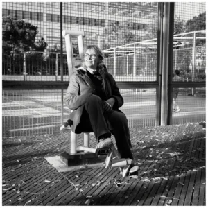 Marc Lavorata · 06/12/2025 · 1103 x 1103 Cette photographie en noir et blanc met en scène une personne assise sur un banc, sujet principal. La composition est simple et épurée, avec un cadrage serré qui isole le sujet des éléments environnants.Le banc est placé sur un sol en bois, et l’arrière-plan montre une clôture métallique et des arbres. La lumière semble naturelle, douce et légèrement contrastée, créant des ombres marquées. L’ambiance est calme et introspective, renforcée par le monochrome. Le style est sobre et documentaire, évoquant une atmosphère contemplative et mélancolique.