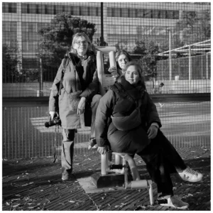 Marc Lavorata · 06/12/2025 · 1103 x 1103 Cette photographie met en scène trois femmes assises sur un banc public. Le sujet principal est leur interaction détendue, avec une composition centrée sur elles.Le cadrage est serré, incluant un arrière-plan urbain flou avec des bâtiments et une clôture. La lumière est naturelle, probablement en journée, créant des contrastes nets entre ombres et clartés.Les couleurs dominantes sont les tons de gris et noir, typiques d’une photo en noir et blanc, accentuant les textures des vêtements et des visages.Le style est documentaire et spontané, évoquant une ambiance calme et authentique.