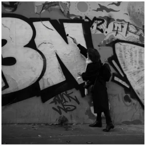 Marc Lavorata · 06/12/2025 · 1103 x 1103 Cette photographie en noir et blanc met en scène un graffeur devant un mur tagué. Le sujet principal est l’artiste, de dos, en train de peindre ou d’observer le mur couvert de graffitis stylisés, dont les lettres "BW" dominent.La composition est simple et centrée, avec un cadrage serré sur le graffeur et le mur. La lumière est douce, créant des contrastes marqués entre ombres et zones éclairées. Les couleurs, bien que monochromes, jouent sur les nuances de gris pour accentuer les détails et les textures.L’ambiance est urbaine, artistique et légèrement mystérieuse, reflétant la culture street art.