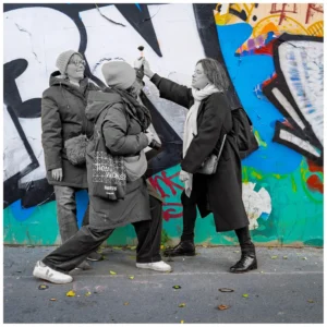 Marc Lavorata · 06/12/2025 · 1103 x 1103 Cette photographie montre un groupe de trois personnes en mouvement devant un mur tagué. Le sujet principal semble être l’interaction entre eux, avec une dynamique de groupe.La composition est dynamique, avec un cadrage serré et des lignes de fuite créées par les postures. La lumière est naturelle, légèrement contrastée, mettant en valeur les contours.Les couleurs dominantes sont vives : bleu, vert, rouge et noir du graffiti, contrastant avec les tons neutres des vêtements. Le style est urbain et spontané, évoquant une ambiance jeune et énergique, presque théâtrale.