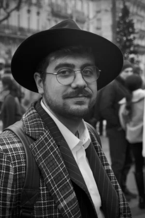 Thomas · 13/12/2025 · 2048 x 3080 Cette photographie en noir et blanc met en scène un homme comme sujet principal, vêtu d’un veston à carreaux, d’une chemise blanche et d’un large chapeau. Le cadrage est serré, centré sur lui, avec un arrière-plan flou suggérant une foule.La lumière semble naturelle, douce, et met en valeur les traits du visage. L’ambiance est sobre et intemporelle, renforcée par le monochrome. Le style évoque une touche documentaire ou artistique, avec une attention portée sur les détails vestimentaires et l’expression faciale. L’atmosphère est à la fois sérieuse et légèrement nostalgique.