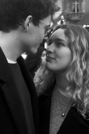 Thomas · 13/12/2025 · 2048 x 3080 Cette photographie en noir et blanc met en scène deux personnes en portrait serré. Le sujet principal est un couple dont les visages sont proches, créant une intimité. Le cadrage est serré, centré sur leurs têtes et épaules. La lumière douce et naturelle accentue les contrastes faciaux, avec des ombres marquées.L’ambiance est romantique et chaleureuse, renforcée par le style sobre et élégant des vêtements. Les tons dominants, bien que monochromes, varient des gris clairs aux noirs profonds, ajoutant une touche dramatique. L’arrière-plan flou et peu visible maintient l’attention sur les protagonistes.