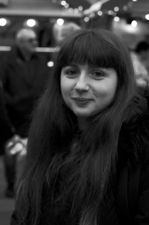 Thomas · 13/12/2025 · 2048 x 3080 Cette photographie en noir et blanc met en valeur une jeune femme comme sujet principal, au premier plan. Son visage est légèrement flou, avec des cheveux longs et une expression neutre. Le cadrage est serré sur elle, bien que l'arrière-plan montre des silhouettes floues, suggérant une foule.La lumière est douce, éclairant principalement son visage. L'absence de couleurs accentue les contrastes et les textures, comme ses cheveux et sa peau. Le style est documentaire et naturel, créant une ambiance intimiste et légèrement mystérieuse dans un contexte probablement urbain ou événementiel.
