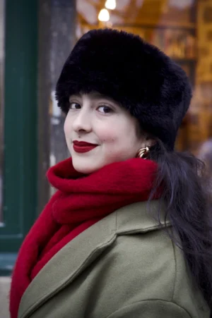 Thomas · 13/12/2025 · 2048 x 3080 Cette photographie met en scène une femme comme sujet principal, souriante et élégante. Le cadrage est serré sur son visage et le haut de son corps, avec un arrière-plan flou et légèrement décentré.La lumière douce et naturelle met en valeur ses traits, créant des ombres douces. Les couleurs dominantes sont les tons chauds : rouge vif du foulard et du lipstick, vert olive de la veste, et noir du chapeau.Le style évoque un mélange rétro et sophistiqué, renforcé par les accessoires vintage. L’ambiance est chaleureuse et intime, évoquant une touche de nostalgie et de charme classique.