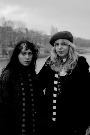 Thomas · 13/12/2025 · 2048 x 3080 Cette photographie en noir et blanc met en scène deux jeunes femmes comme sujets principaux. Elles sont placées côte à côte, l’une avec des cheveux longs et l’autre portant une écharpe et un chapeau. La composition est simple et symétrique, avec un cadrage serré sur elles.La lumière est douce et naturelle, sans ombres marquées. Les couleurs dominantes, bien que monochromes, jouent sur les contrastes de tons clairs et foncés.Le style évoque une ambiance rétro ou vintage, renforcée par les vêtements et coiffures. L’atmosphère est intime et légèrement mélancolique, avec une touche de nostalgie.