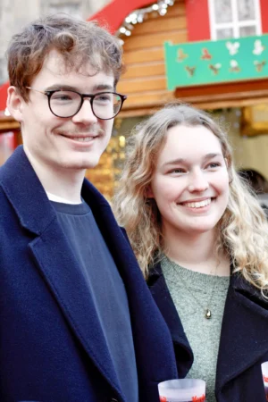 Cédric · 13/12/2025 · 3456 x 5184 Cette photographie montre deux jeunes posant côte à côte. Le sujet principal est un couple souriant, probablement en fête.La composition est serrée, avec un cadrage rapproché mettant en valeur leurs visages. La lumière est naturelle et douce, éclairant bien leurs traits. Les couleurs dominantes sont sobres : tons neutres (bleu marine, vert foncé) et touches chaudes (peau, rouge des décorations en arrière-plan).Le style est simple et spontané, évoquant une ambiance conviviale et festive, probablement lors d’un événement hivernal ou de Noël.