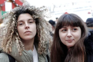 Cédric · 13/12/2025 · 5184 x 3456 Cette photographie met en scène deux jeunes femmes comme sujets principaux. La composition est serrée, les cadrant en portrait côte à côte. Le cadrage est proche, mettant l'accent sur leurs visages et expressions neutres.La lumière semble naturelle, légèrement tamisée, sans ombres marquées. Les couleurs dominantes sont des tons neutres et naturels : peaux claires, cheveux bruns et gris, avec des touches de vert et de rouge en arrière-plan.Le style est sobre et réaliste, évoquant une ambiance quotidienne et spontanée, peut-être urbaine. L'atmosphère est calme et introspective.