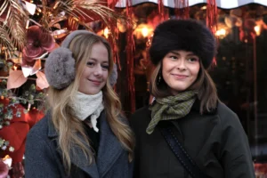 Cédric · 13/12/2025 · 5184 x 3456 Cette photographie met en scène deux femmes souriantes, habillées chaudement pour l’hiver. Le sujet principal est leur complicité et leur tenue festive.La composition est symétrique, avec les deux femmes côte à côte. Le cadrage est serré, centré sur elles, avec un arrière-plan flou mais évocateur de décorations de Noël.La lumière est douce et naturelle, éclairant leurs visages. Les couleurs dominantes sont les tons froids (gris, noir) et chauds (rouge des décorations, beige des accessoires).Le style est naturel et chaleureux, évoquant une ambiance conviviale et hivernale.