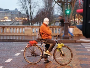 Cédric · 20/12/2025 · 4361 x 3270 Cette photographie met en scène un homme âgé comme sujet principal, assis sur un vélo près d’un pont. La composition montre une scène urbaine paisible, avec des éléments architecturaux et des arbres en arrière-plan.Le cadrage est serré sur le sujet, tout en intégrant l’environnement. La lumière, douce et tamisée, suggère un moment crépusculaire ou hivernal. Les couleurs dominantes sont les tons neutres (gris, beige) et les touches chaudes (orange du vêtement, jaune des fleurs).Le style est documentaire et naturel, capturant une ambiance calme et mélancolique, typique d’une ville en hiver.