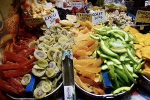 Cédric · 20/12/2025 · 5184 x 3456 Cette photographie montre un étal de marché riche en produits frais. Le sujet principal est une variété de légumes et fruits, comme des avocats, des asperges, des champignons et des radis, disposés sur des plateaux.La composition est dynamique, avec un cadrage serré mettant en valeur les couleurs vives. Les tons dominants sont verts, jaunes et blancs, avec des touches de rouge et de brun. La lumière, naturelle et douce, accentue les textures et les couleurs, créant une ambiance chaleureuse et vivante, typique d’un marché local. Le style est réaliste et photographique, sans retouche apparente.