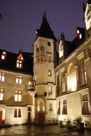 Cédric · 20/12/2025 · 3388 x 5082 Cette photographie met en scène un **château médiéval** éclairé la nuit. Le **sujet principal** est une tour centrale avec une horloge, entourée de bâtiments de style gothique.La **composition** est symétrique, avec des lignes verticales accentuant la hauteur des structures. Le **cadrage** est serré, centré sur la tour.Les **lumières chaudes** des fenêtres créent un contraste avec l’obscurité ambiante, mettant en valeur les détails architecturaux. Les **couleurs dominantes** sont des tons beiges et gris, adoucis par les éclairages dorés.L’ambiance est **mystérieuse et majestueuse**, évoquant une époque historique. Le style est **classique et monumental**.