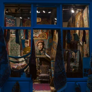 Don · 3245 x 3245 Cette photographie montre une vitrine remplie d’objets textiles et décoratifs colorés. Le sujet principal semble être une collection d’artisanat, incluant des tissus brodés, des vêtements traditionnels et des portraits encadrés.La composition est dense, avec un cadrage serré qui met en valeur les détails. Les couleurs dominantes sont vives et chaudes : bleus profonds, ors, rouges et motifs ethniques. La lumière, tamisée et artificielle, crée une ambiance chaleureuse et intimiste, évoquant un style traditionnel et artisanal. L’ensemble dégage une atmosphère riche et culturelle.