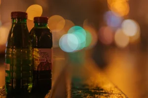 Don · 6476 x 4317 Cette photographie met en scène deux bouteilles, dont une verte et une brune, posées sur une surface floue. Le sujet principal est ces bouteilles, probablement de boisson.La composition est simple et centrée, avec un cadrage serré. L'arrière-plan est flou, créant un effet bokeh avec des lumières colorées, ce qui attire l'attention sur les bouteilles.Les couleurs dominantes sont les verts et bruns des bouteilles, contrastant avec les teintes chaudes et lumineuses de l'arrière-plan. Le style est épuré et artistique, évoquant une ambiance chaleureuse et conviviale, typique d'un bar ou d'un restaurant la nuit.