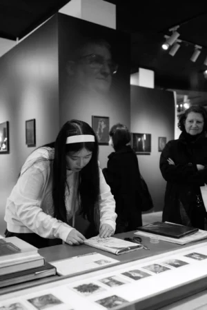 Philippe · 2001 x 3000 Cette photographie en noir et blanc montre une jeune femme, sujet principal, portant des lunettes futuristes et examinant des documents sur une table. La composition met en valeur un espace intérieur, probablement une exposition ou une bibliothèque.Le cadrage est serré sur elle, avec des reflets et des personnes en arrière-plan légèrement flous. La lumière est douce et uniforme, créant des contrastes modérés. L’ambiance est studieuse et technologique, renforcée par les éléments rétro-futuristes. Le style est documentaire, avec une touche artistique due aux jeux de lumière et aux reflets.